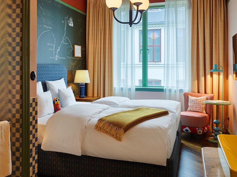 Eclectic hotel guest room with a green-painted window frame, a cozy pink armchair, and a bed set against a dark chalkboard wall with hand-drawn maps.