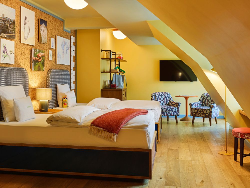 Spacious attic hotel family room showing wood floors, two beds set against a cork accent wall, sloped yellow ceilings, and a cozy seating area under a skylight window.