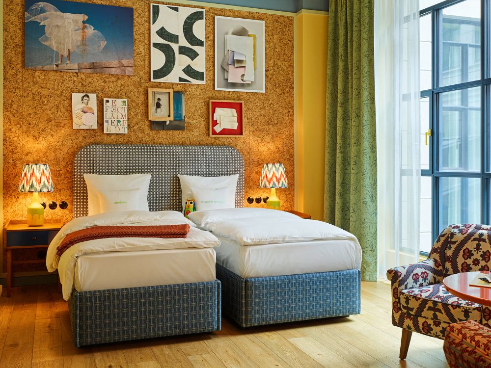 Boutique hotel twin bedroom featuring two beds side-by-side with patterned headboards, set against a cork accent wall decorated with framed gallery art.