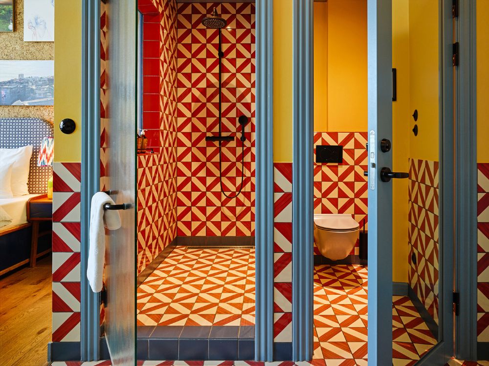 Vibrant hotel bathroom showcasing a walk-in shower and toilet area decorated in matching red and white triangular patterned tiles with blue trim.