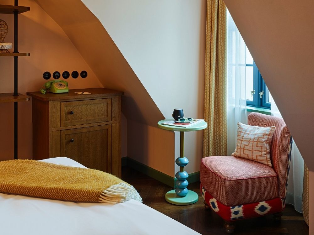 Quirky hotel attic suite corner featuring a red patterned armchair, a fun turquoise pedestal side table, and a wooden chest with a retro green phone under a sloped ceiling.