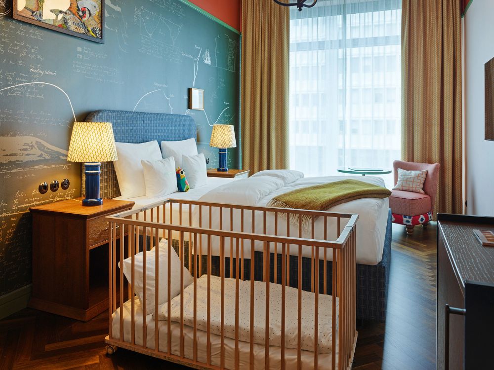Family-friendly hotel bedroom featuring a double bed under blue bedside lamps, a chalkboard wall, and a wooden baby crib in the foreground.