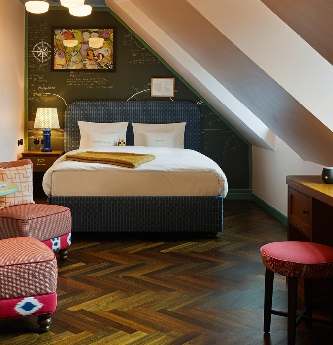 Quirky attic hotel suite with a sloped ceiling, rich dark wood herringbone parquet flooring, a blue bed, and colorful upholstered armchairs.