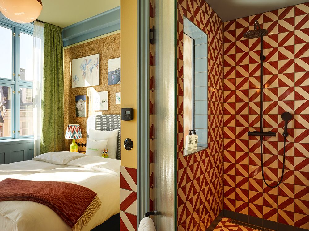 View from a boutique hotel bedroom with a cork-paneled wall looking into a bright walk-in shower tiled in red and white triangles.