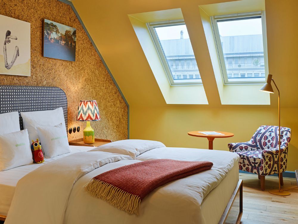 Cosy hotel attic bedroom under two angled skylight windows featuring a yellow accent wall, a cork headboard, and a patterned fabric armchair.