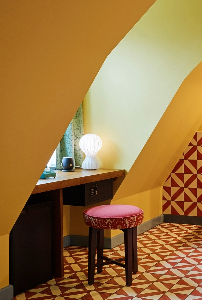 Cozy hotel attic writing desk nestled under a slanted yellow eave, featuring a glowing white mushroom lamp, a pink stool, and bold geometric floor tiles.