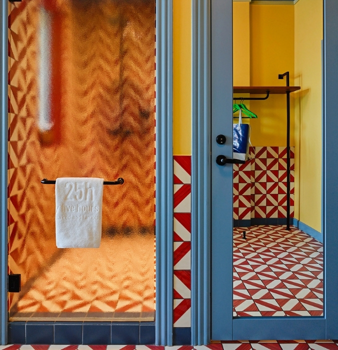 Frosted glass hotel shower door with a branded 25hours white towel, reflecting bold red and white geometric wall tiles.