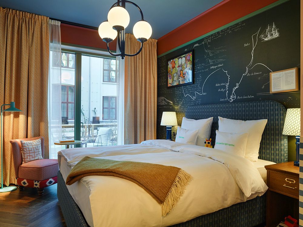 Spacious hotel bedroom featuring a dark green chalkboard accent wall decorated with chalk illustrations, a blue bed, and a red armchair.