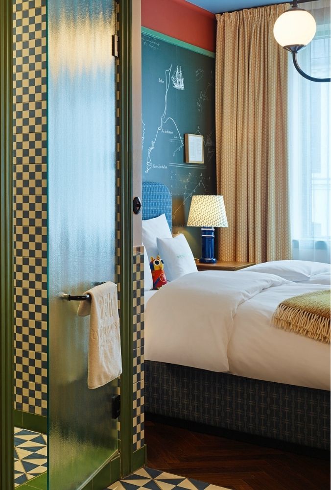 View past a blue-and-white checkered bathroom tile wall into an eclectic hotel bedroom featuring a dark green chalkboard wall and a blue bed.
