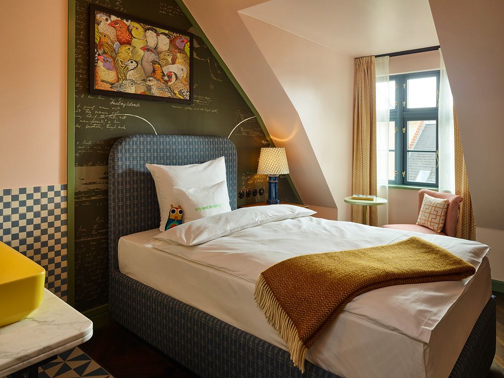 Quirky attic hotel bedroom featuring a sloped white ceiling, a plush bed with a blue headboard, a chalkboard accent wall, and a framed painting of birds.
