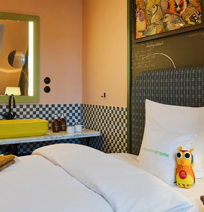 Plush hotel bed with a yellow owl plush toy next to an open-concept washbasin vanity featuring blue and white checkered tiles.