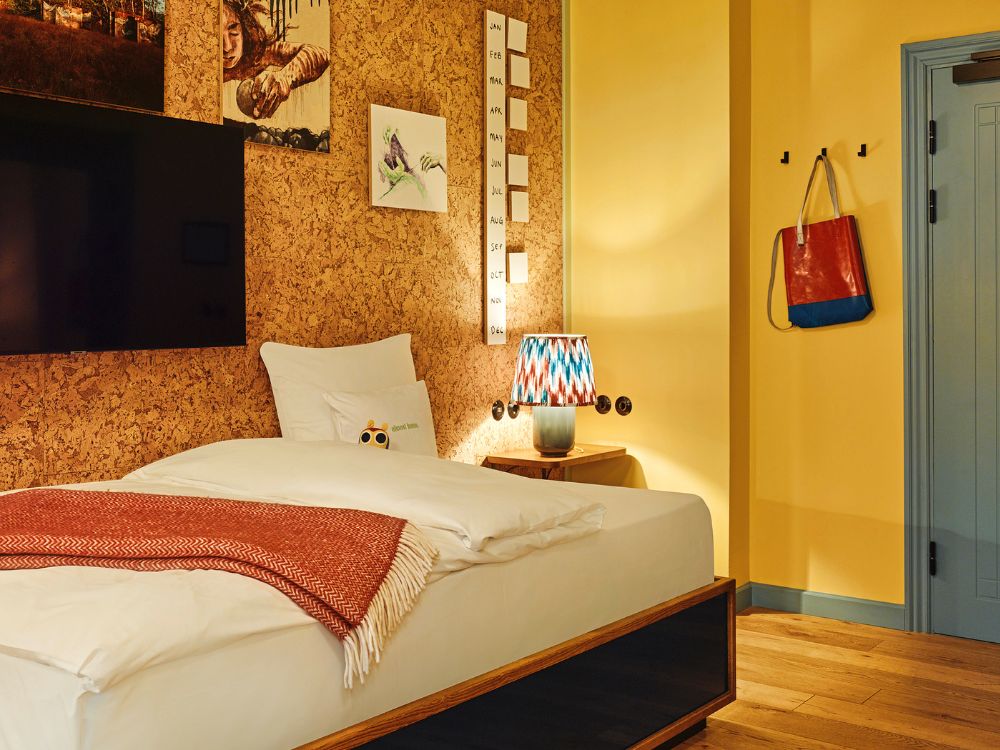 Modern hotel room bed set against a cork-paneled wall with framed artwork, a colorful patterned table lamp, and a red tote bag hanging on a yellow wall.