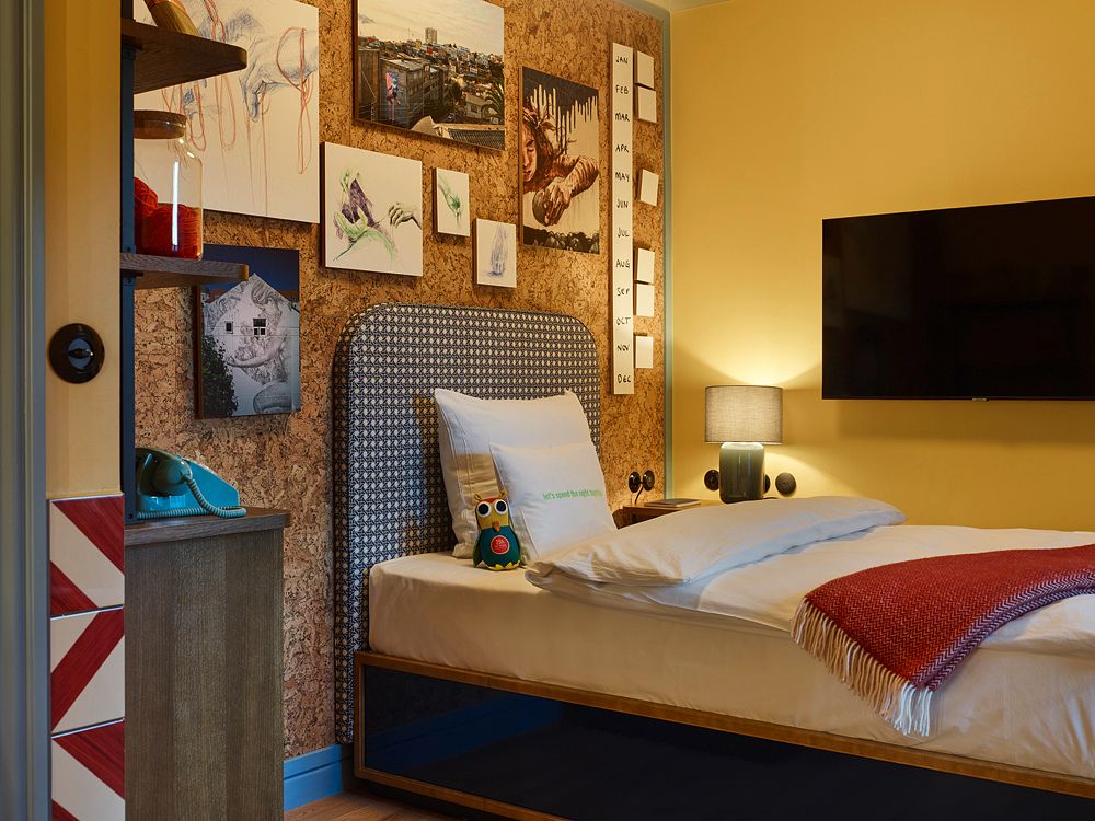 Eclectic hotel bedroom showing a bed against a cork headboard wall, a wall-mounted flat-screen TV, and a retro turquoise rotary phone on a desk.
