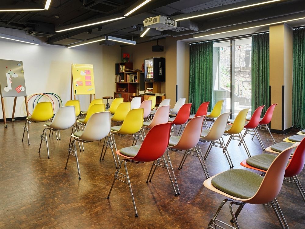 A room with rows of yellow, red, and gray chairs facing a screen, whiteboard, and bookshelf, with green curtains over large windows.