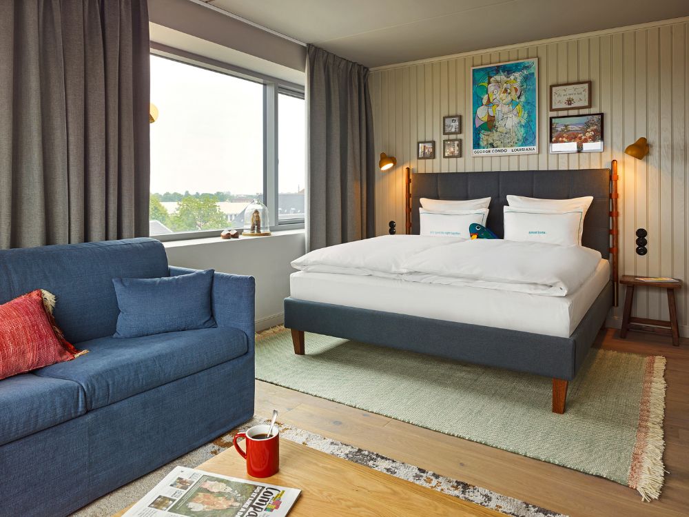 Spacious hotel guest room featuring a plush double bed on a green rug, a comfortable blue sofa with a red pillow, and large windows.