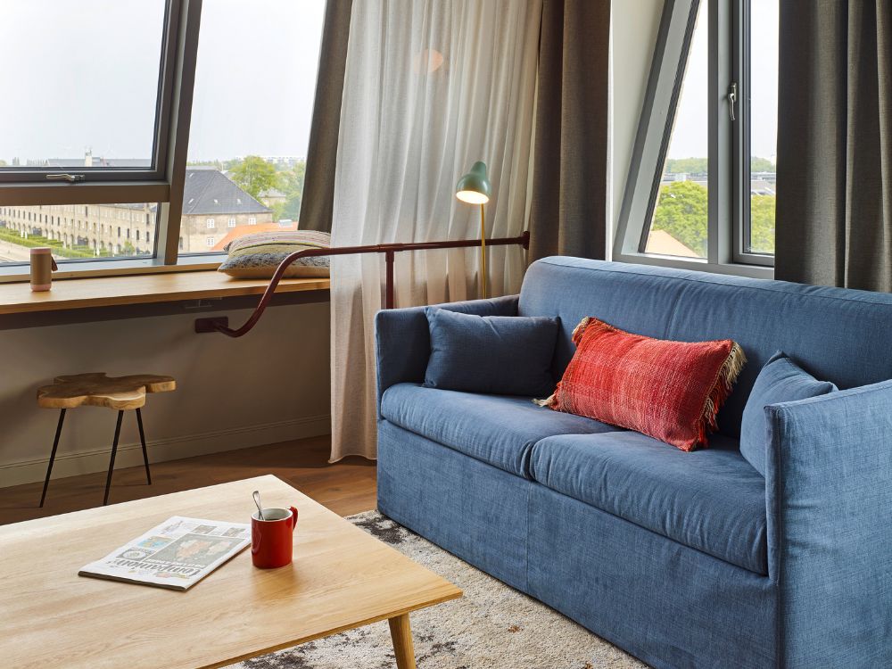 Comfortable blue sofa with a red accent pillow next to a wide wooden window sill overlooking a scenic city waterfront.