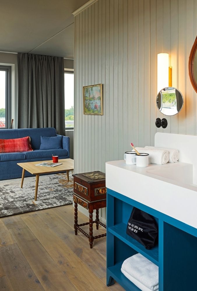 Open-plan hotel bathroom vanity with a white sink on a blue cabinet looking through to a cozy blue sofa sitting area with a vintage chest.
