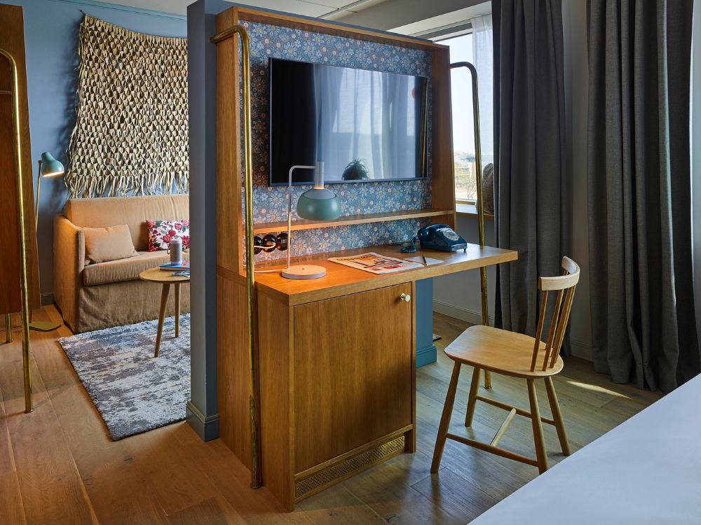 Boutique hotel desk integrated into a wooden partition wall featuring a built-in TV with floral wallpaper backing and a wooden chair.
