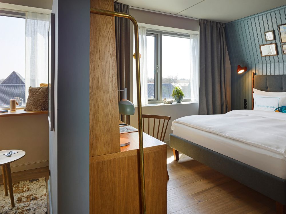 Modern hotel guest room featuring a wooden desk partition dividing a plush double bed and a cozy window sitting area.