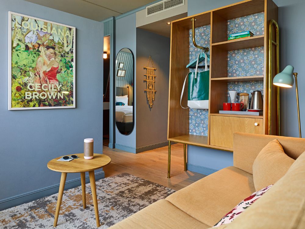 Cozy hotel suite living room featuring a beige sofa, a floral-backed wooden wardrobe unit, and a colorful art poster on a blue wall.