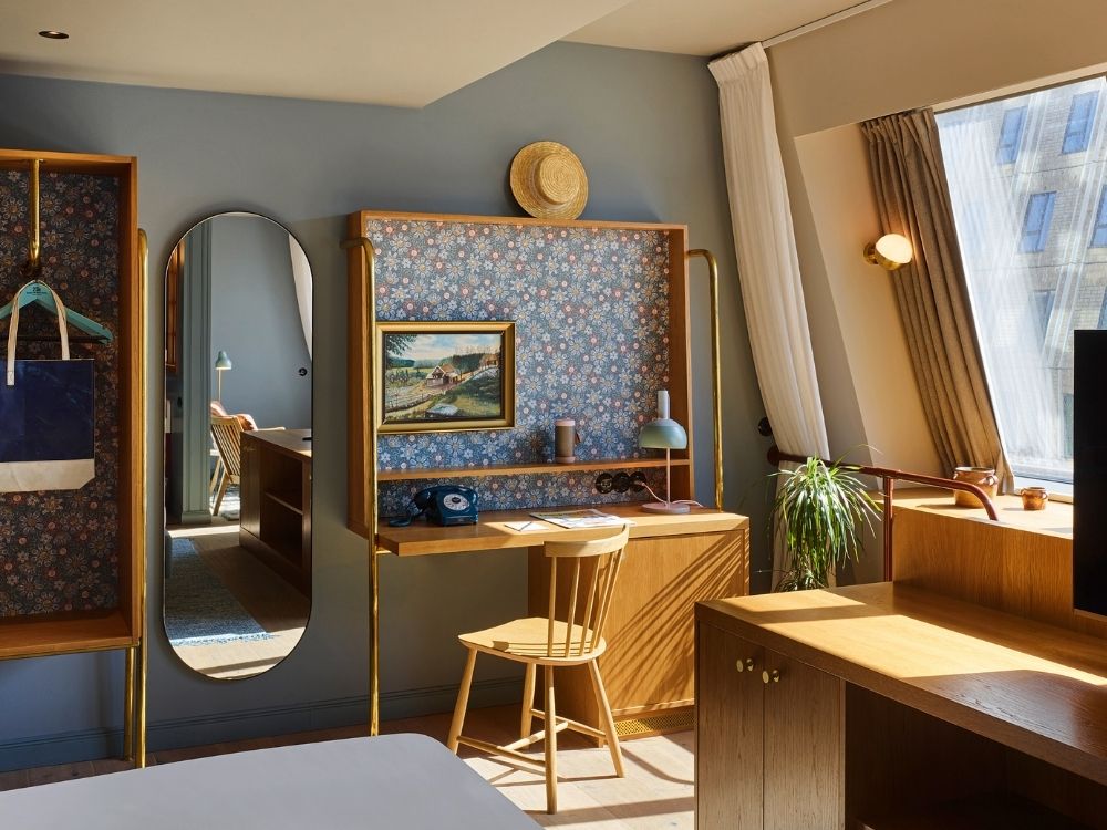 Boutique hotel suite featuring an oval wall mirror, a wooden study desk with floral wallpaper backing, a wardrobe rack, and a bed next to a bright slanted window.