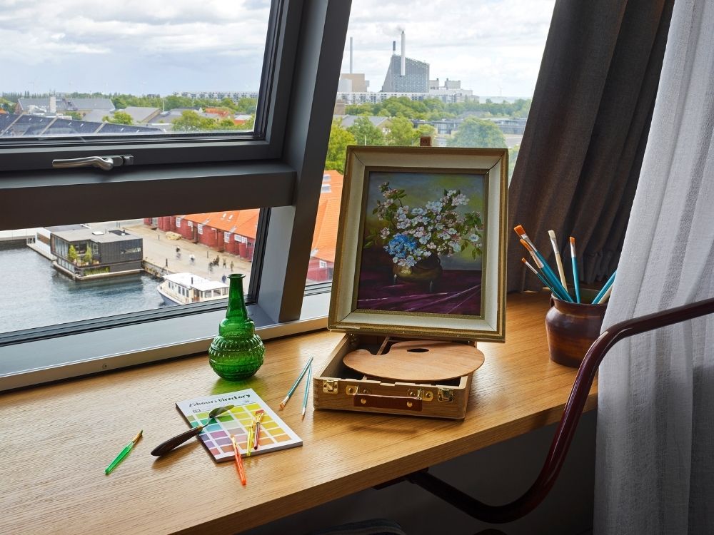 An artist's painting easel with a floral oil painting, paintbrushes, and color swatches arranged on a wide wooden window sill overlooking a scenic harbor.