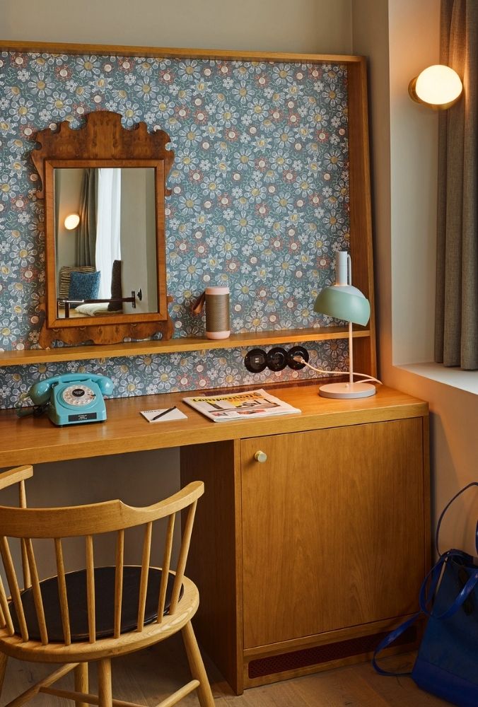 Chic hotel room desk workspace featuring a wooden writing desk, an ornate wood-framed wall mirror, blue floral wallpaper, a green lamp, and a retro blue phone.