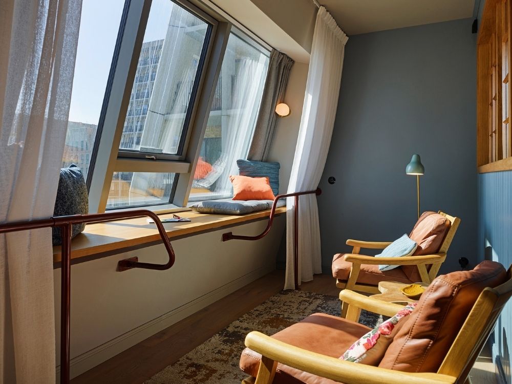 Relaxing hotel lounge corner featuring two brown leather armchairs, a wooden stool, and a long wooden window seat under slanted skylight windows.
