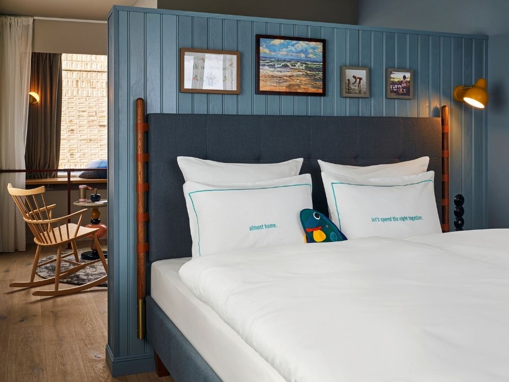 Plush hotel bed with a grey button-tufted headboard against a blue wood-paneled partition wall, with a glimpse of a cozy rocking chair seating area behind.