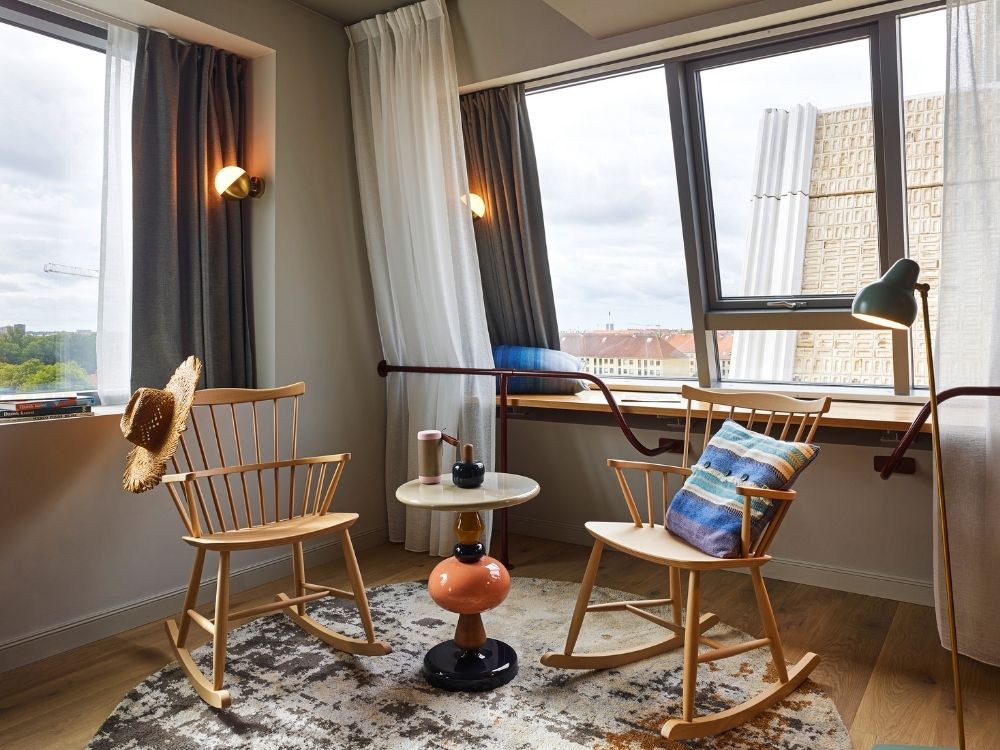 Chic seating area in a boutique hotel room featuring two light wood rocking chairs, a colorful pedestal side table, and a printed rug next to a large window.