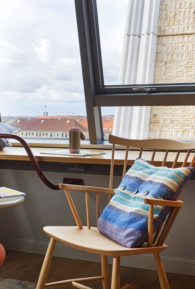 A light wood spindle chair with a blue striped knit pillow sitting by a sunny window ledge overlooking a scenic city view.