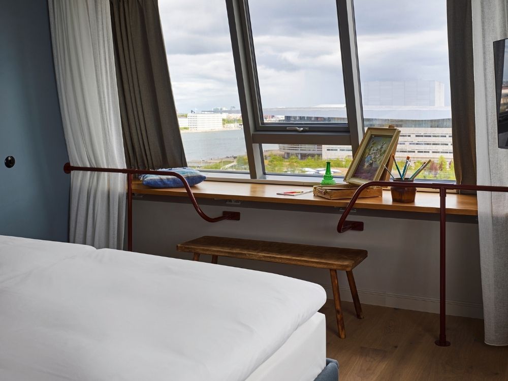 Wide wooden window seat used as an artist's station with an easel, paintbrushes, and a rustic wooden bench overlooking a city canal and modern opera house.