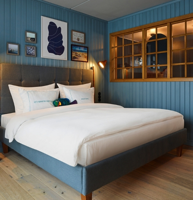 Boutique hotel bed with a grey button-tufted headboard against blue vertical wood paneling, featuring a decorative internal wood-framed window grid.