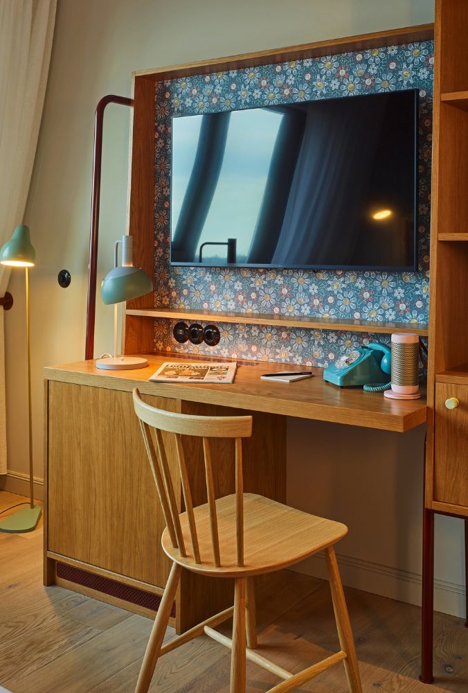 Compact hotel study desk featuring a wooden chair, a retro blue phone, and a flat-screen TV mounted against a floral wallpaper backdrop.