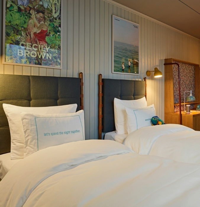 Close-up of comfortable twin beds with grey headboards against light vertical wood-paneled walls, decorated with colorful modern posters and reading lamps.