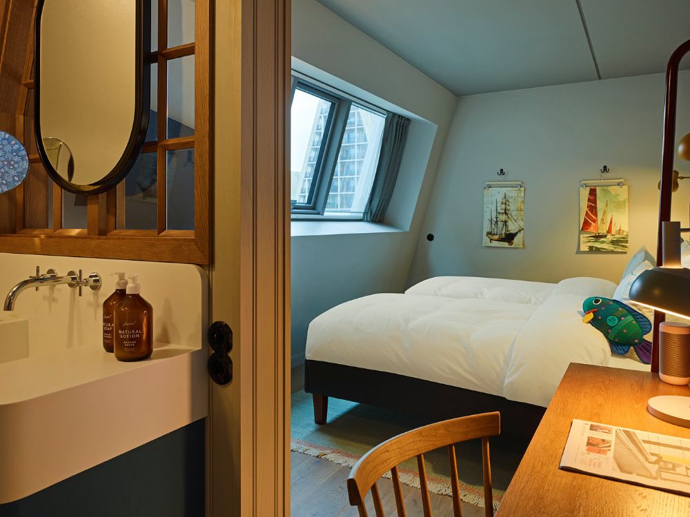 View past an open-concept washroom station with a round mirror into a slanted attic bedroom featuring twin beds and retro travel posters.