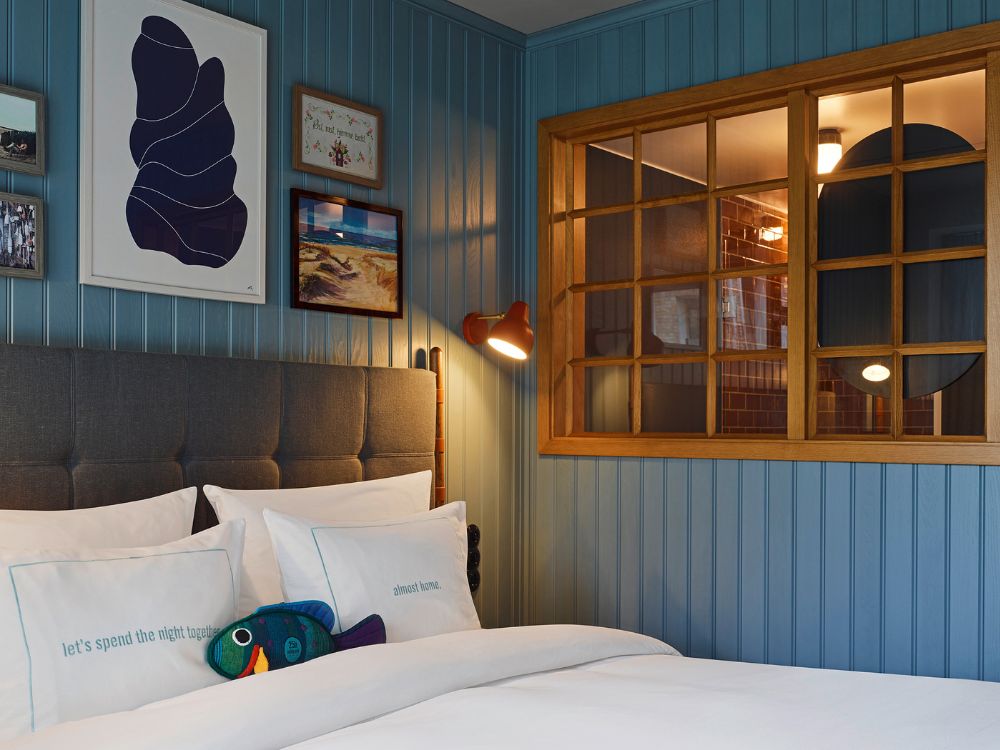 Boutique hotel bed with a grey button-tufted headboard against blue vertical wood paneling, featuring a decorative internal wood-framed window grid.