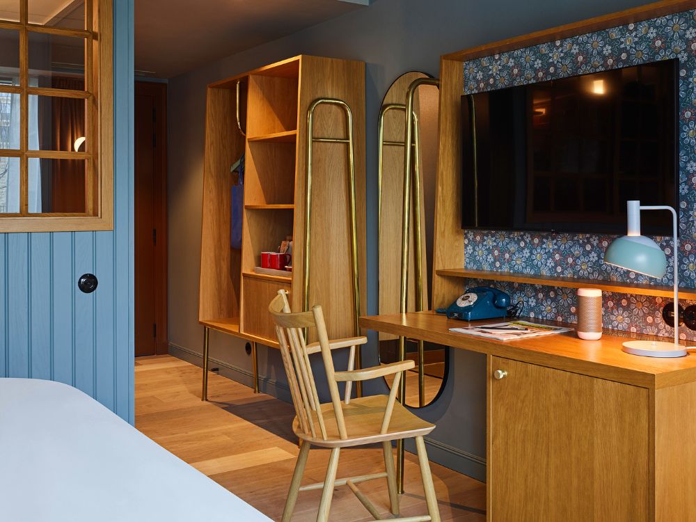 Hotel room study area with a wooden writing desk under a wall-mounted flat-screen TV backed by floral wallpaper, alongside a wooden wardrobe and spindle chair.
