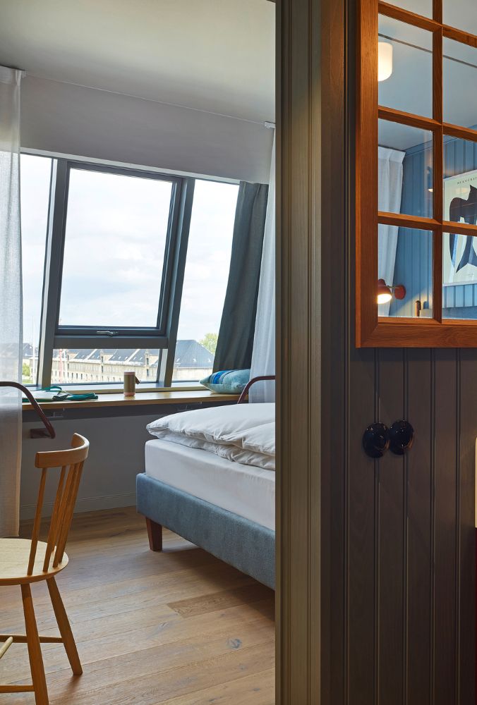 View from a wood-paneled doorway into a modern guest room showing a plush bed with white linens and a wide slanted window.