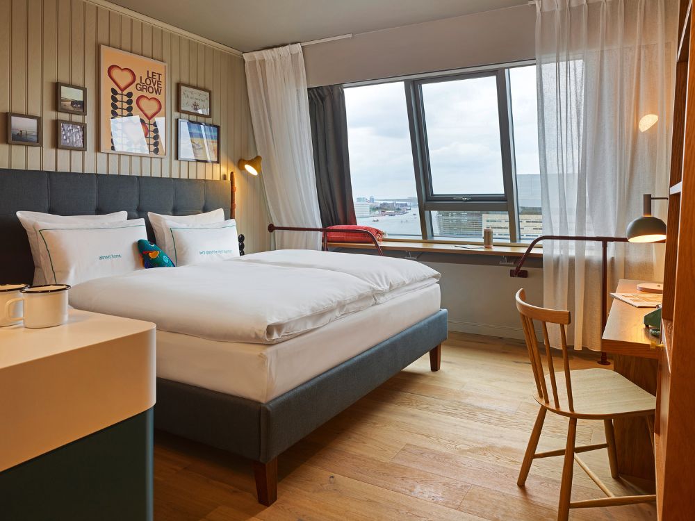 Full boutique hotel bedroom featuring wood floors, a double bed by a wide window, a wooden writing desk with a spindle chair, and colorful wall posters.