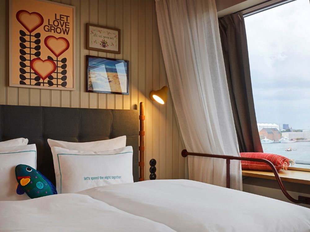 Plush hotel bed next to a wide window overlooking a city harbor, featuring a red throw cushion on the sill and a retro yellow reading lamp.