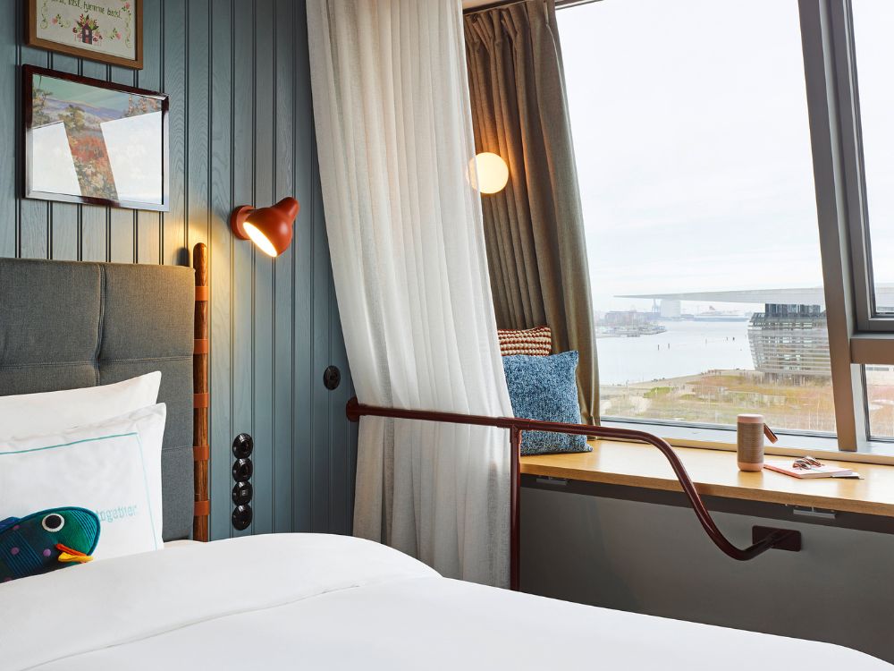 Boutique hotel bed next to a wide wooden window sill used as a bench seat, offering scenic views over a city harbor.