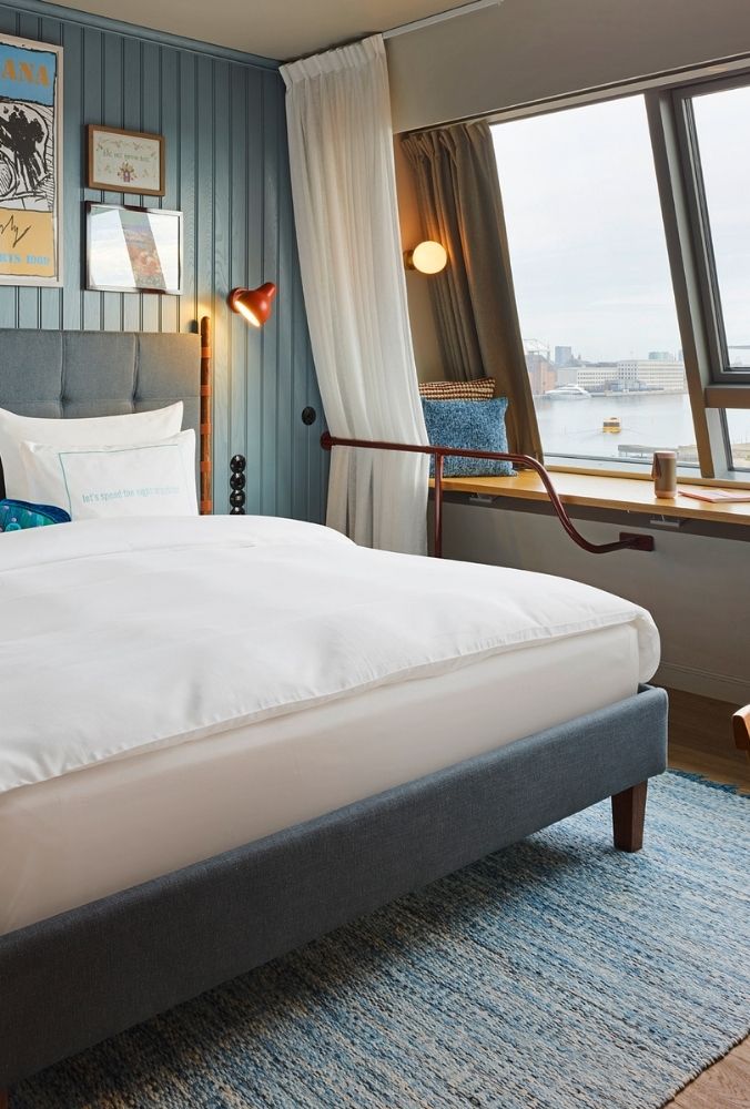 Comfortable double bed on a blue woven rug next to a wide window seat overlooking a city harbor with boats under a cloudy sky.