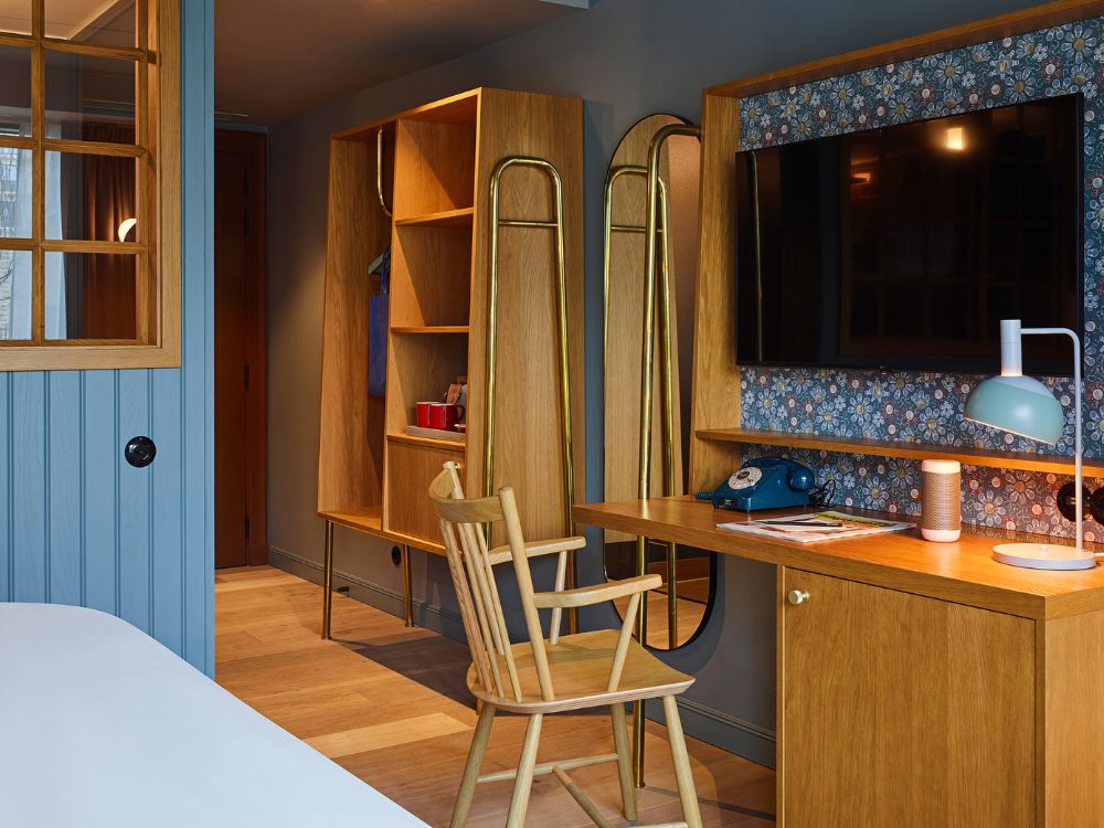 Chic guest room study area featuring a wooden writing desk, a retro blue phone, a green lamp, and a wall-mounted TV backed by floral wallpaper.