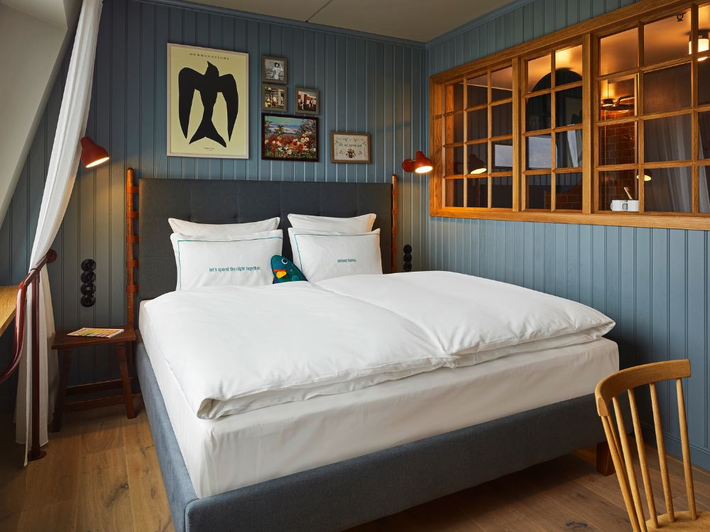 Hotel king bed with a grey tufted headboard set against blue vertical wood-paneled walls featuring a decorative wooden internal window grid.