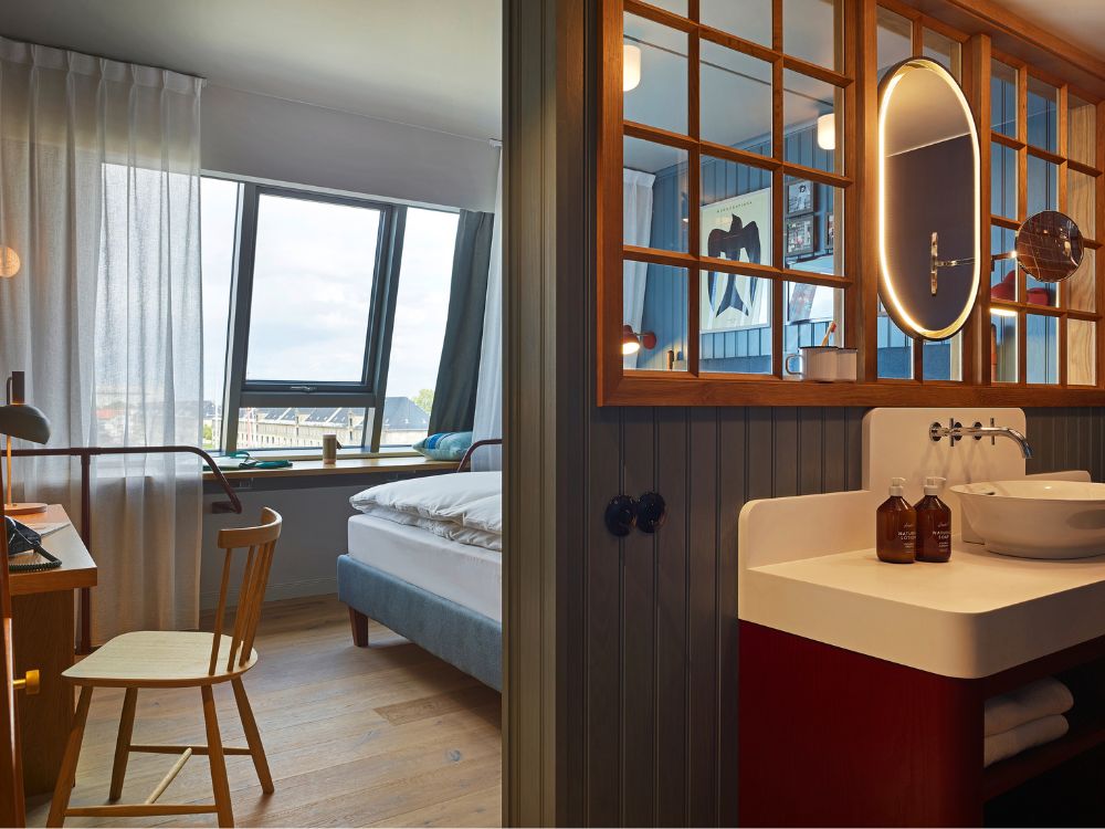 View past an open-concept washbasin with a backlit oval mirror looking into a cozy bedroom featuring a wooden desk chair and a bed by a slanted window.