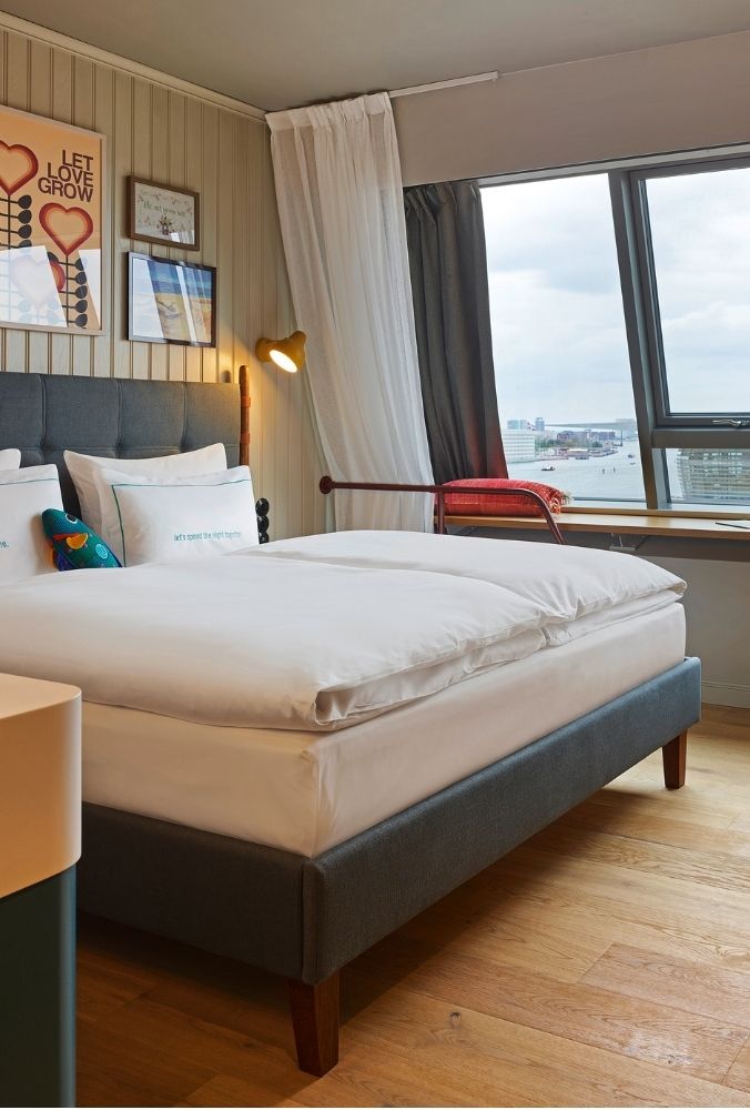 Plush hotel double bed with white duvets next to a wide wooden window seat offering panoramic waterfront views of the city harbor.