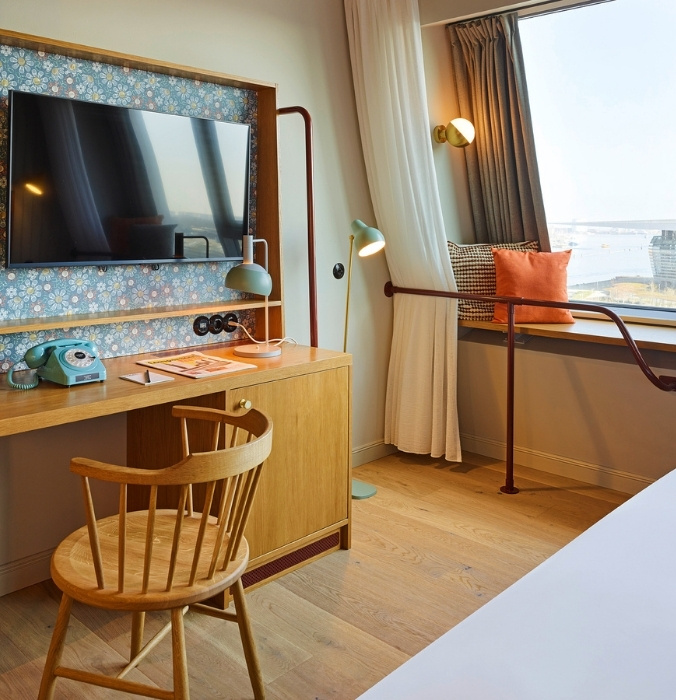 Compact hotel study desk with a wooden chair, a retro blue rotary telephone, and a wall-mounted flat-screen TV on floral wallpaper next to a window seat.