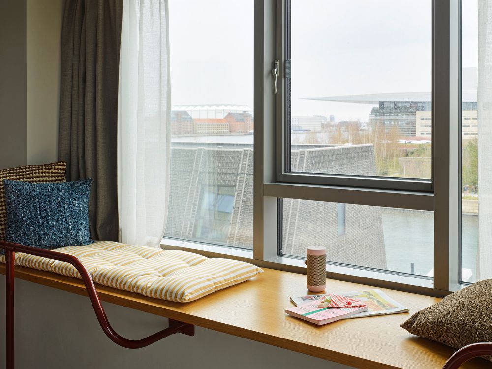 Cozy hotel window bench seat with a yellow-and-white striped cushion, blue and brown accent pillows, and a scenic city view.