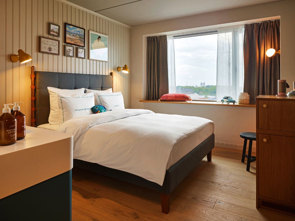 Cozy hotel double room featuring light vertical wood-paneled walls, framed artwork, a plush bed with white linens, and a wide window ledge overlooking the city.